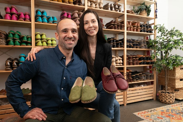The image features two individuals in a shoe store, each holding a pair of shoes. One is holding green shoes, the other maroon, both pairs resembling suede. Shelves behind them display various colorful shoes, including boots, loafers, and sandals. The wooden shelves are multi-leveled, and a patterned rug lies on the floor. A potted plant adds greenery to the scene.