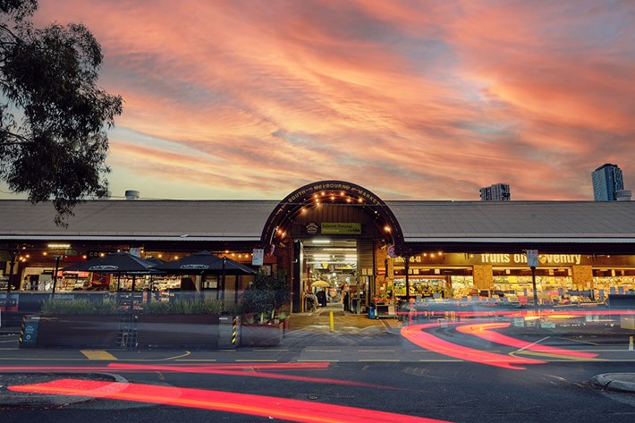 The image captures the entrance of South Melbourne Market at dusk, with the sky displaying pink and orange sunrise hues. The market’s entrance features a prominent archway with the name ‘South Melbourne Market’ at the top. Below, various stalls are visible under the market’s shelter, illuminated by warm lights. The foreground shows light trails from moving vehicles, creating vibrant red streaks along the street. The bustling atmosphere of the historic marketplace is evident as night transitions to day.