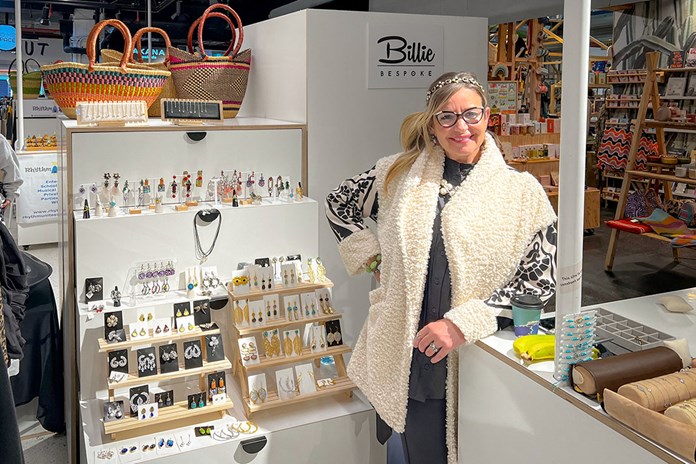 A person stands in front of a display of various jewelry items in a retail store. The display includes earrings, necklaces, and bracelets arranged on white stands and shelves. To the left, there are colorful woven baskets on the top shelf. In the background, there are other products and displays with signage reading “Billie Bespoke.” The setting appears to be an indoor market or boutique shop with a focus on handcrafted goods.