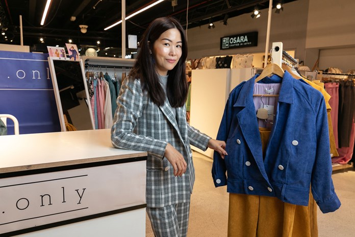 The image features a person in the SO ME Space at South Melbourne Market, examining a blue jacket on a mannequin. The individual is wearing a checkered suit and the mannequin is dressed in a yellow dress beneath the jacket. Various clothing items are displayed in the background, along with store signs “The Only” and “OSARA.” The setting is lit with artificial lighting, typical of retail spaces