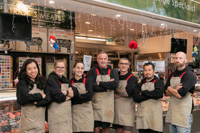 The image features a group of seven people wearing aprons, standing in front of a meat counter at “KIRKPATRICK’S MEATS” butcher shop. The counter displays various meats, and the shop is adorned with hanging lights and informational signs about different cuts of beef and lamb.