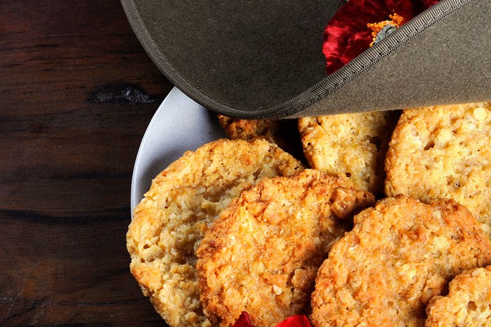 The image features a plate of golden brown, ANZAC biscuits partially obscured by a grey hat with a red poppy reminiscent of ANZAC Day, all resting on a dark wooden surface. The biscuits have a rough, crunchy texture and the felt material of the hat is soft to the touch. The scene is devoid of any logos or text, presenting a simple yet intriguing composition of food and fashion.