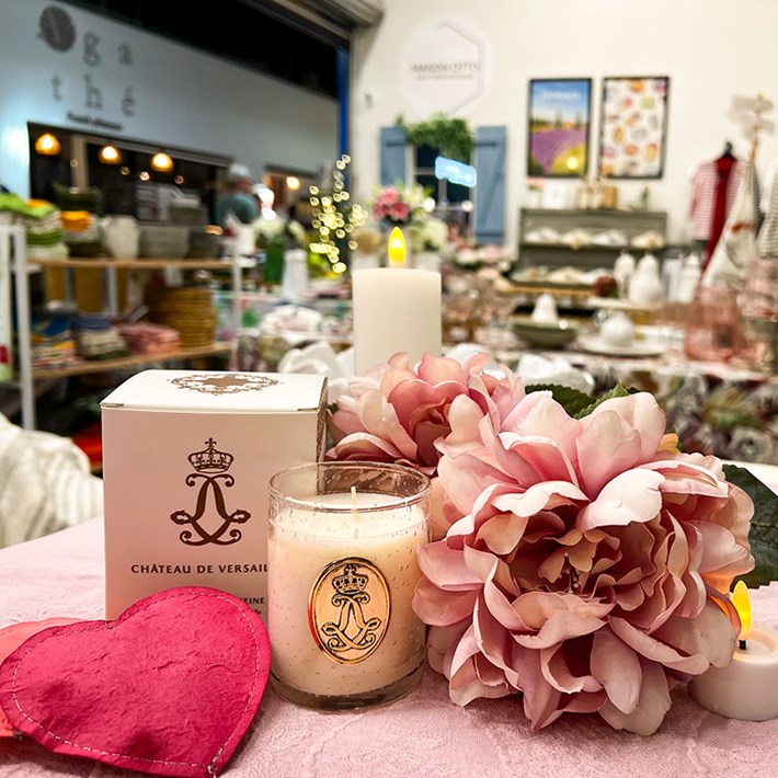 The image features a charming boutique store interior with a pink table at the forefront displaying a white box labeled “CHATEAU DE VERSAILLES”, a lit candle, and a bouquet of pink flowers, creating a warm and inviting atmosphere. The background is filled with various items for sale, including clothing and decorative pieces, all bathed in soft lighting.